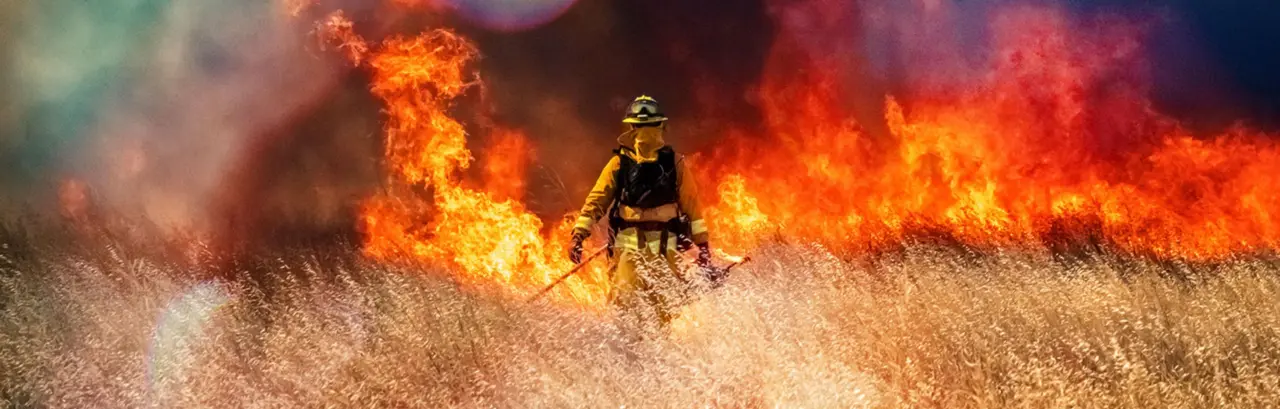 Wildfire raging across grass meadow in California, firefighter walking through meadow