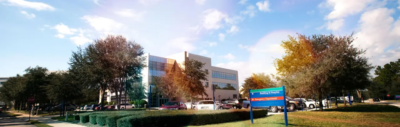 Modern building and parking lot under a partly cloudy sky. Lush trees surround the area.