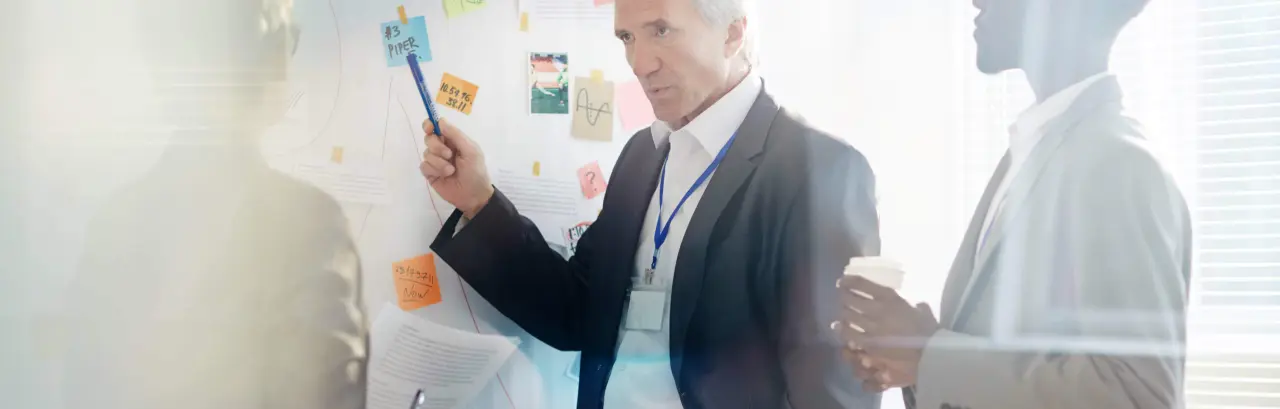 An older man is pointing to a sticky note on a wall with his pen in a group meeting room