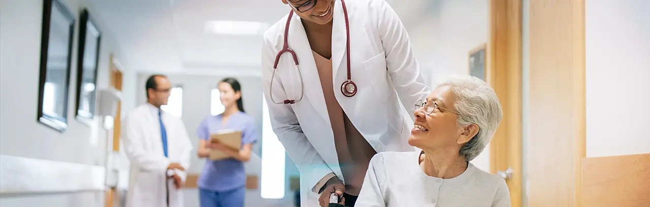 A female doctor pushing a senior patient on a wheelchair in a hospital, smiling at each other