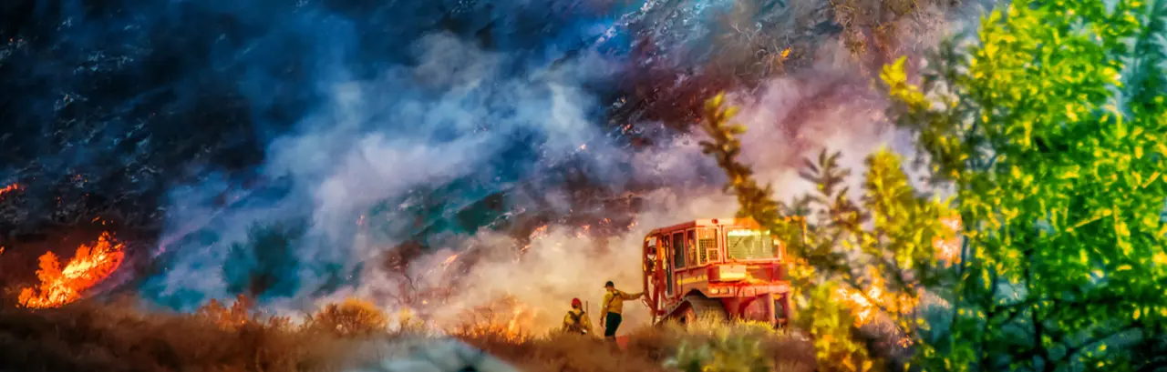 Two firefighters stand alongside bulldozer in California Woolsey Fire with hill burning behind them.