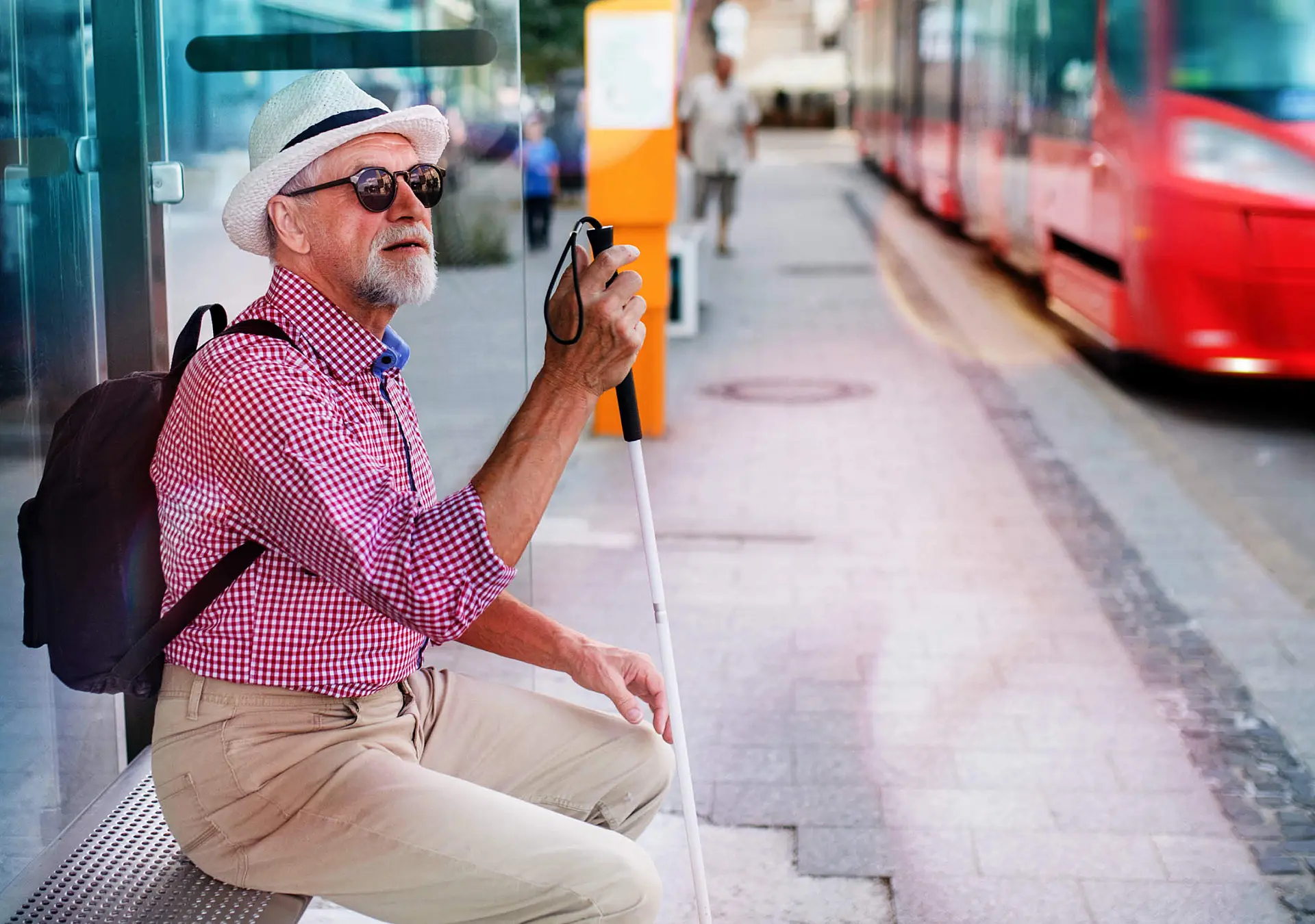 Senior blind man sitting on bench with white cane waiting for public transport in city