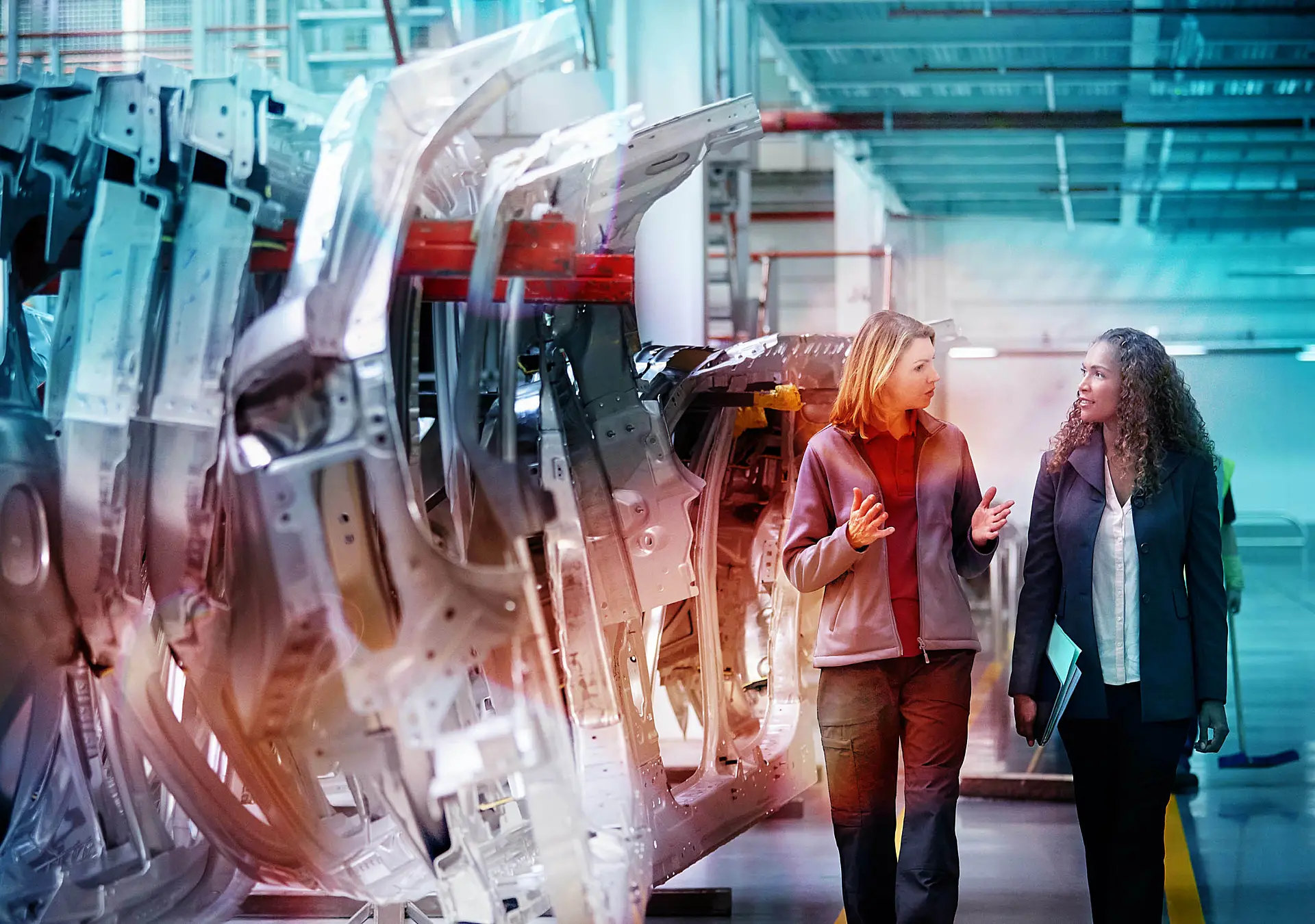 Female engineers discussing by car chassis walking on an aisle in a factory. 