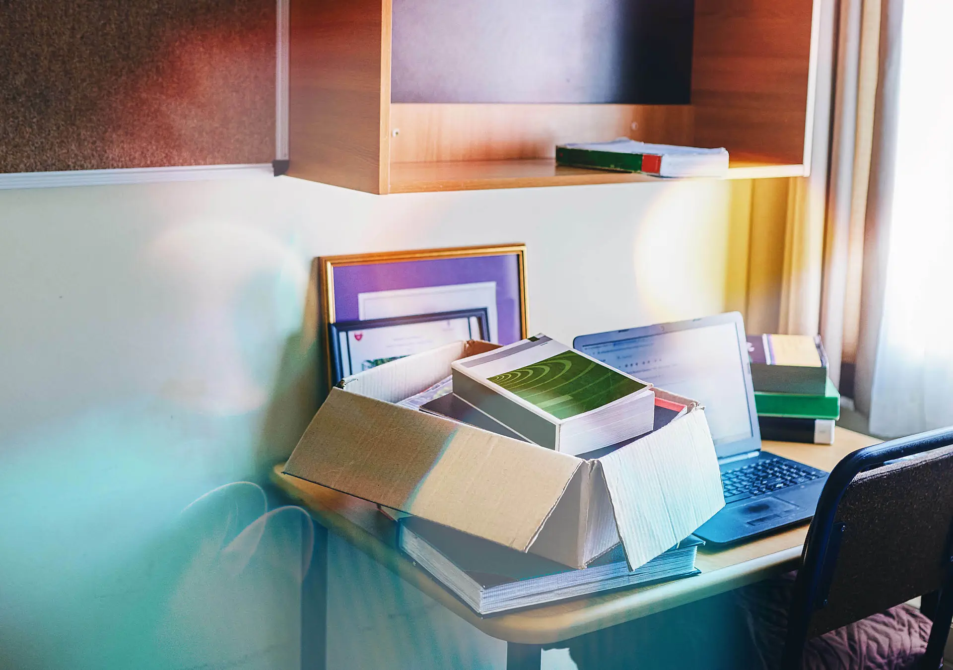 Small desk in a room with a box of books and personal items in it next to a computer