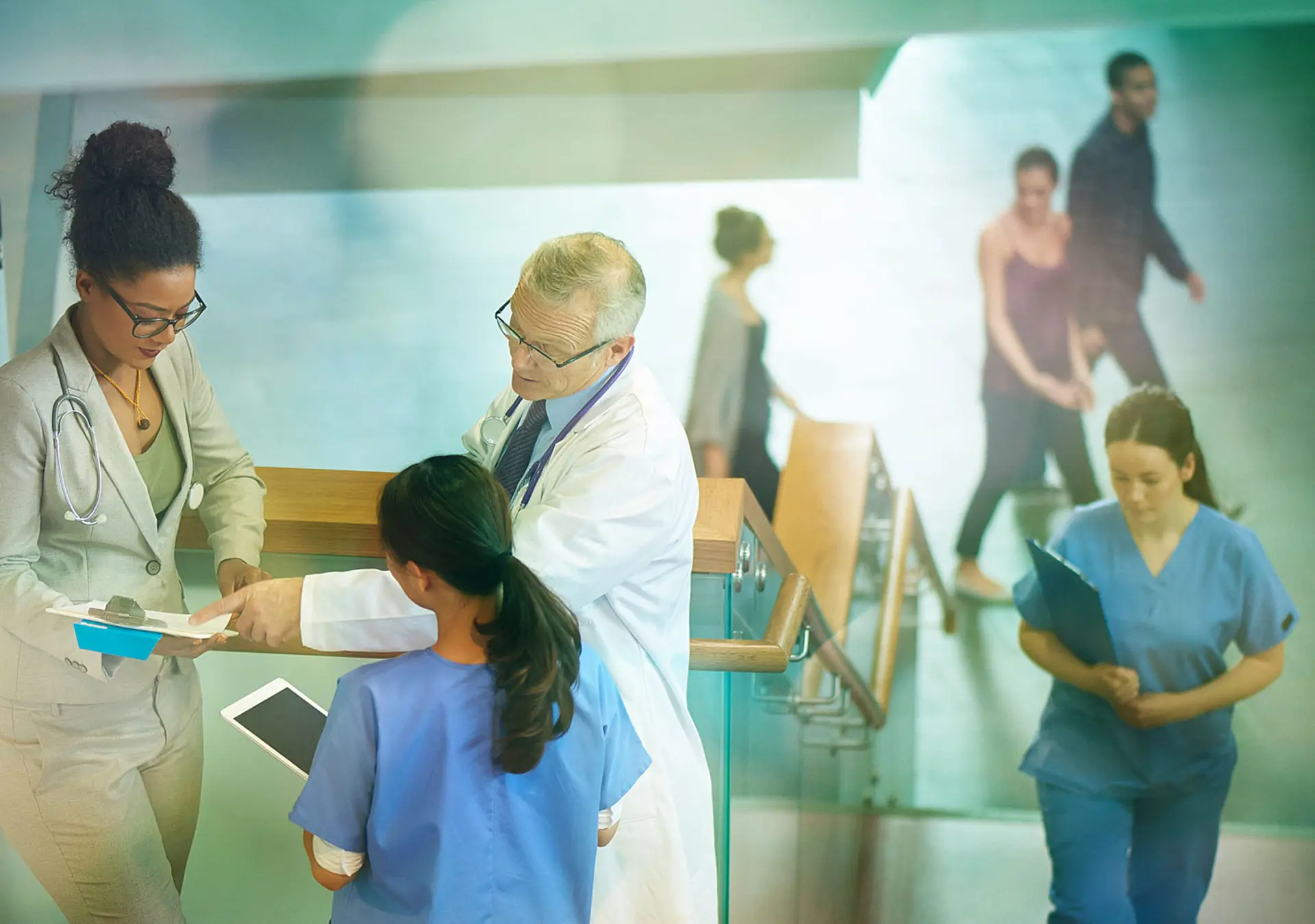 Medical professionals stand on a stairwell of a modern hospital and discuss some case notes.