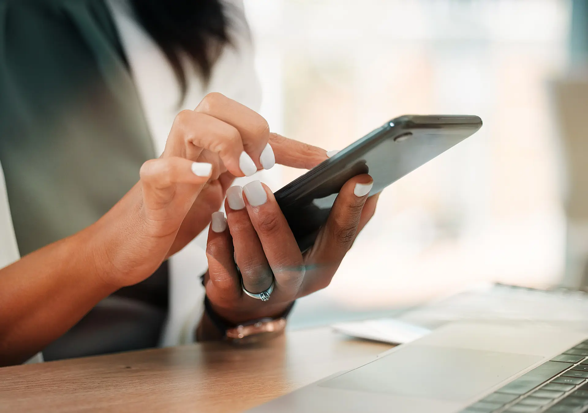 woman at desk with laptop, holding and using her smartphone