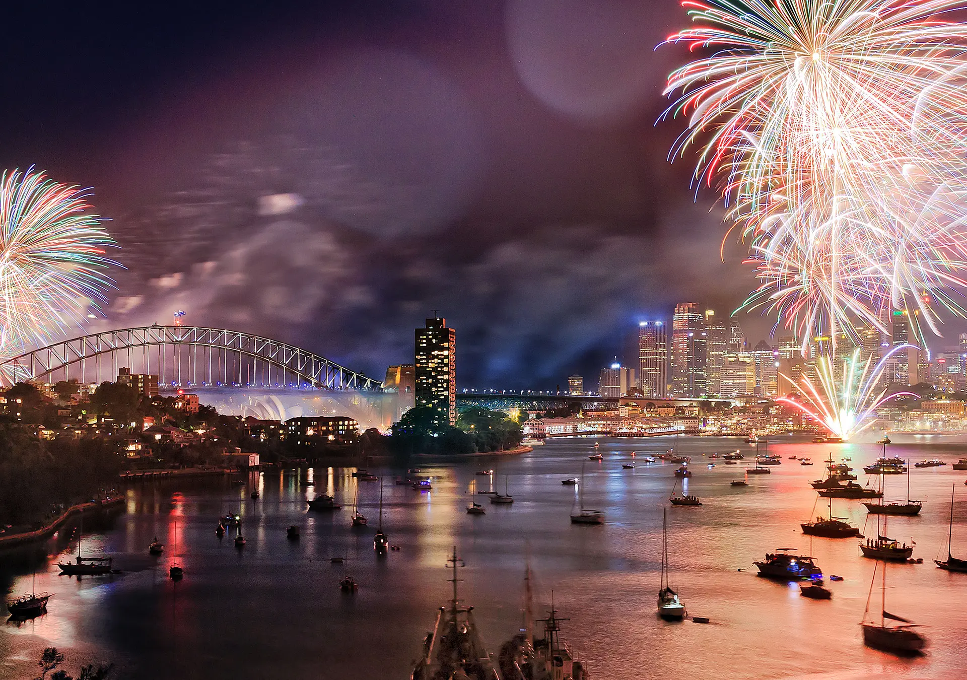 Sydney New Year eve fireworks over Harbour with bridge and city CBD buildings reflecting in water.