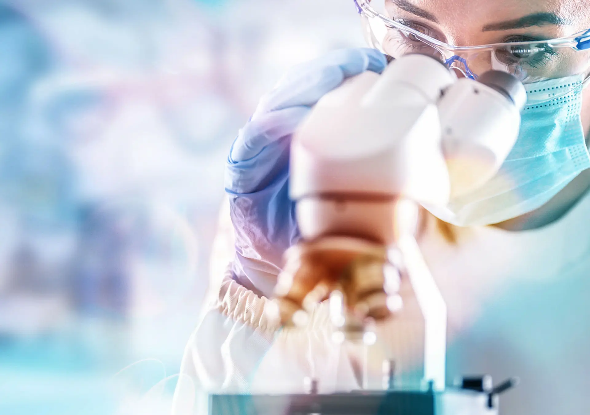 Female lab technician in protective glasses, gloves and face mask next to a microscope in a lab.
