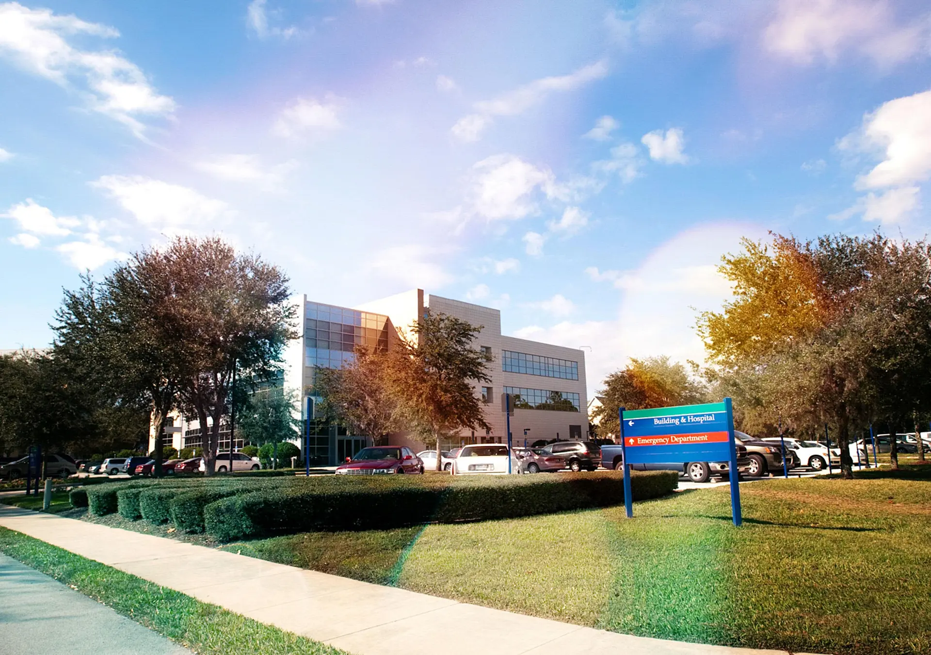Modern building and parking lot under a partly cloudy sky. Lush trees surround the area.