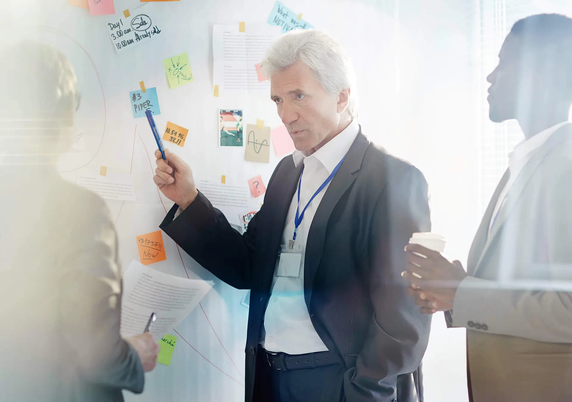 An older man is pointing to a sticky note on a wall with his pen in a group meeting room