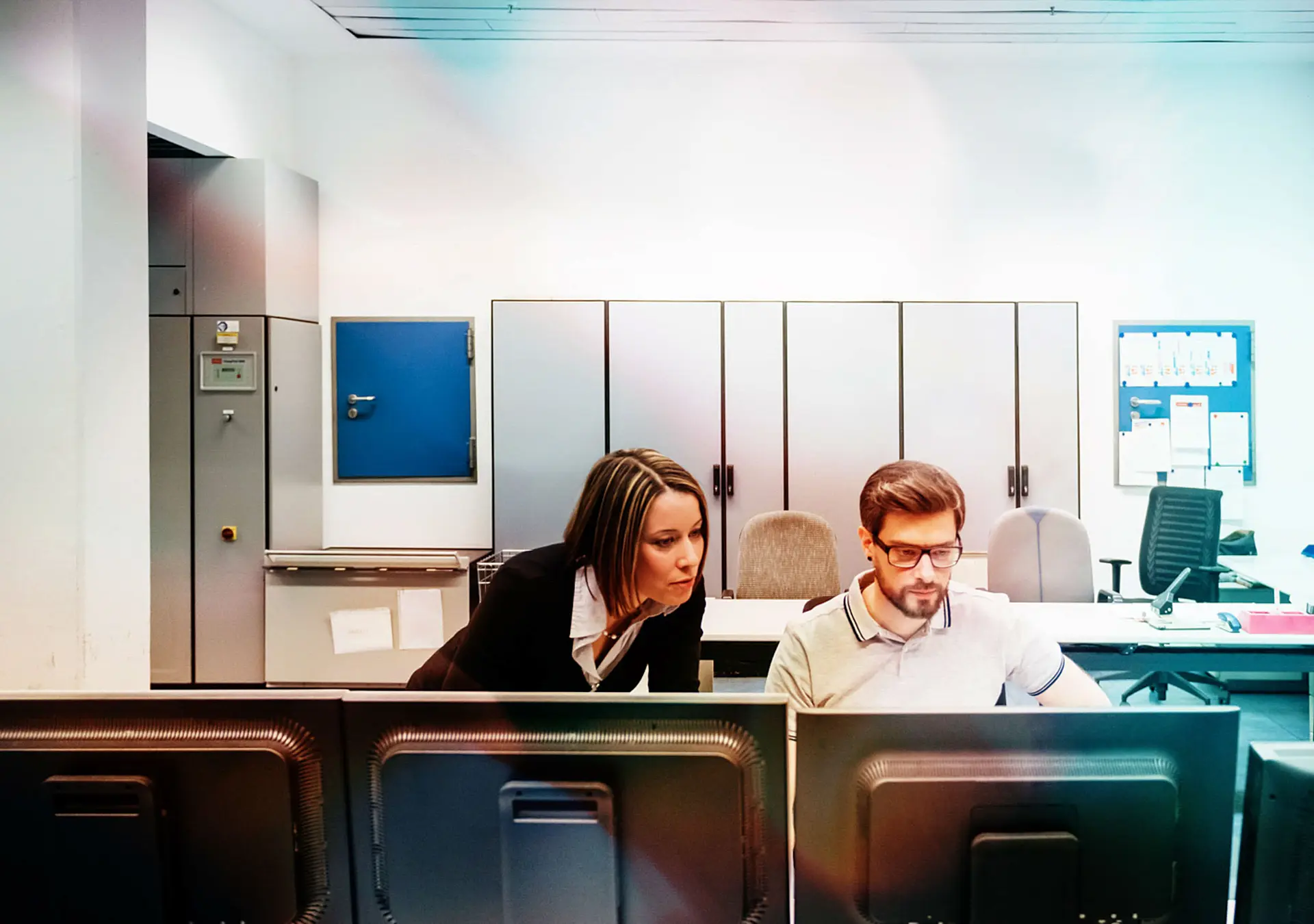 A operations manager assisting her colleague working at multiple computers in the control room.