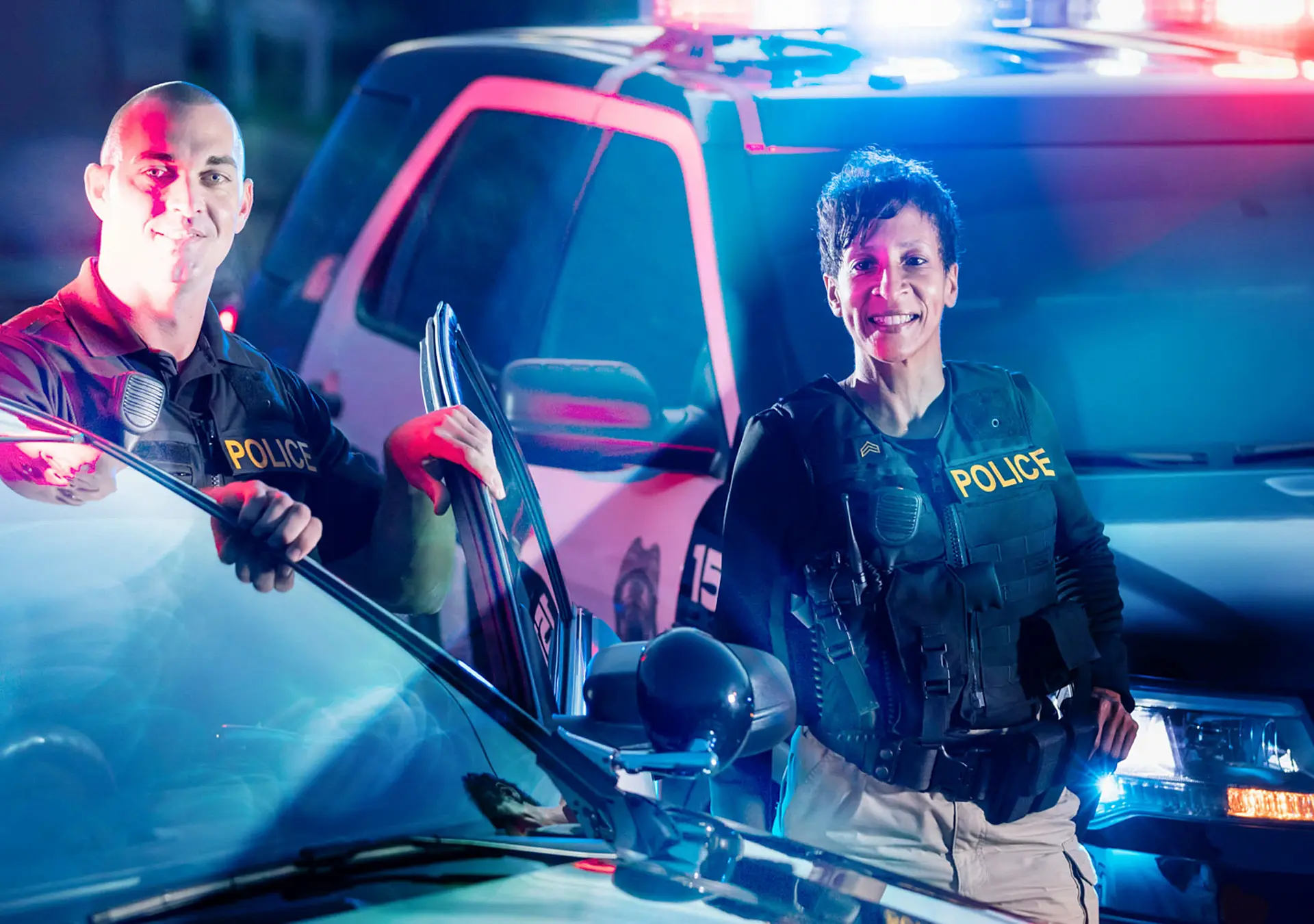 Two police officers standing by the patrol cars, wearing bulletproof vests and duty belts.