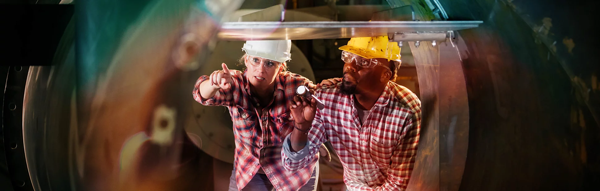 Two people with hard hats examine the inside of a machinery, one pointing, one holds a flashlight.