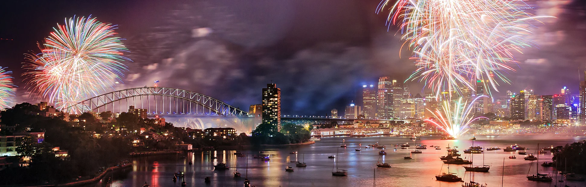 Sydney New Year eve fireworks over Harbour with bridge and city CBD buildings reflecting in water.