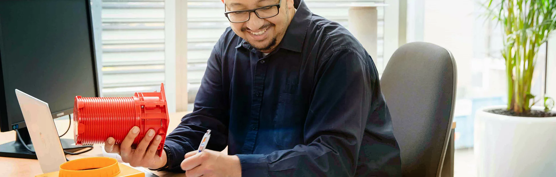 Male engineer with glasses holding a piece of fire protection equipment in his office at his desk.