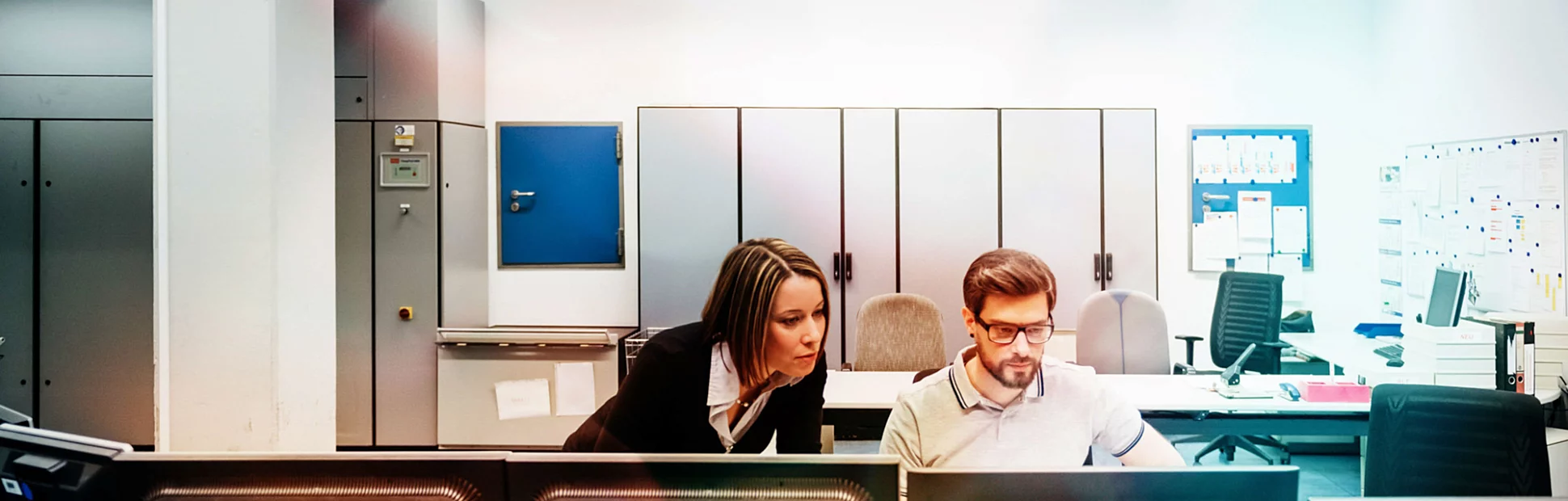 A operations manager assisting her colleague working at multiple computers in the control room.
