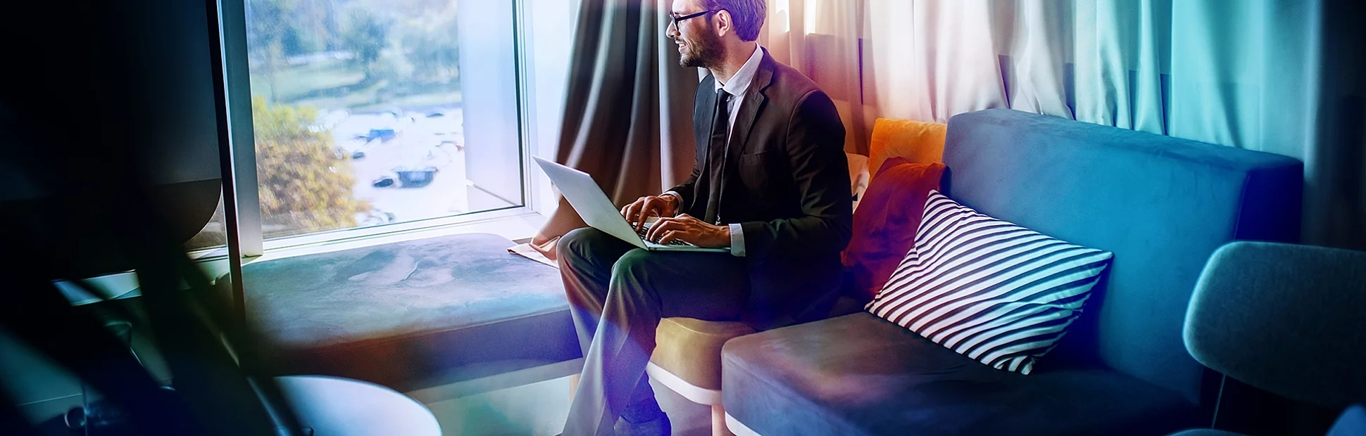 businessman in suit and with eyeglasses sitting in office on sofa, holding laptop in lap
