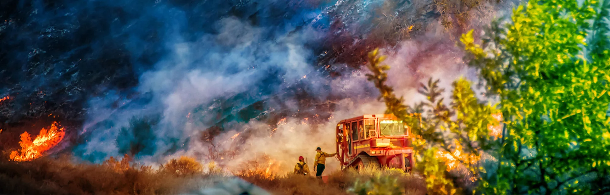 Two firefighters stand alongside bulldozer in California Woolsey Fire with hill burning behind them.