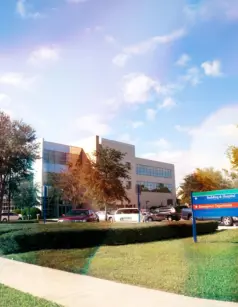 Modern building and parking lot under a partly cloudy sky. Lush trees surround the area.
