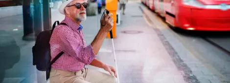 Senior blind man sitting on bench with white cane waiting for public transport in city