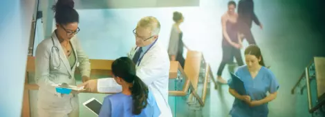 Medical professionals stand on a stairwell of a modern hospital and discuss some case notes.