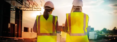 Two women, both in hard hats, walk side by side in front of a building