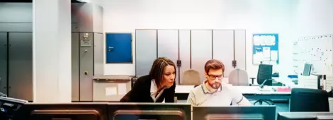 A operations manager assisting her colleague working at multiple computers in the control room.