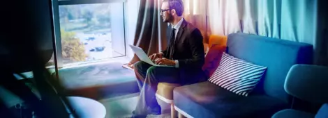 businessman in suit and with eyeglasses sitting in office on sofa, holding laptop in lap