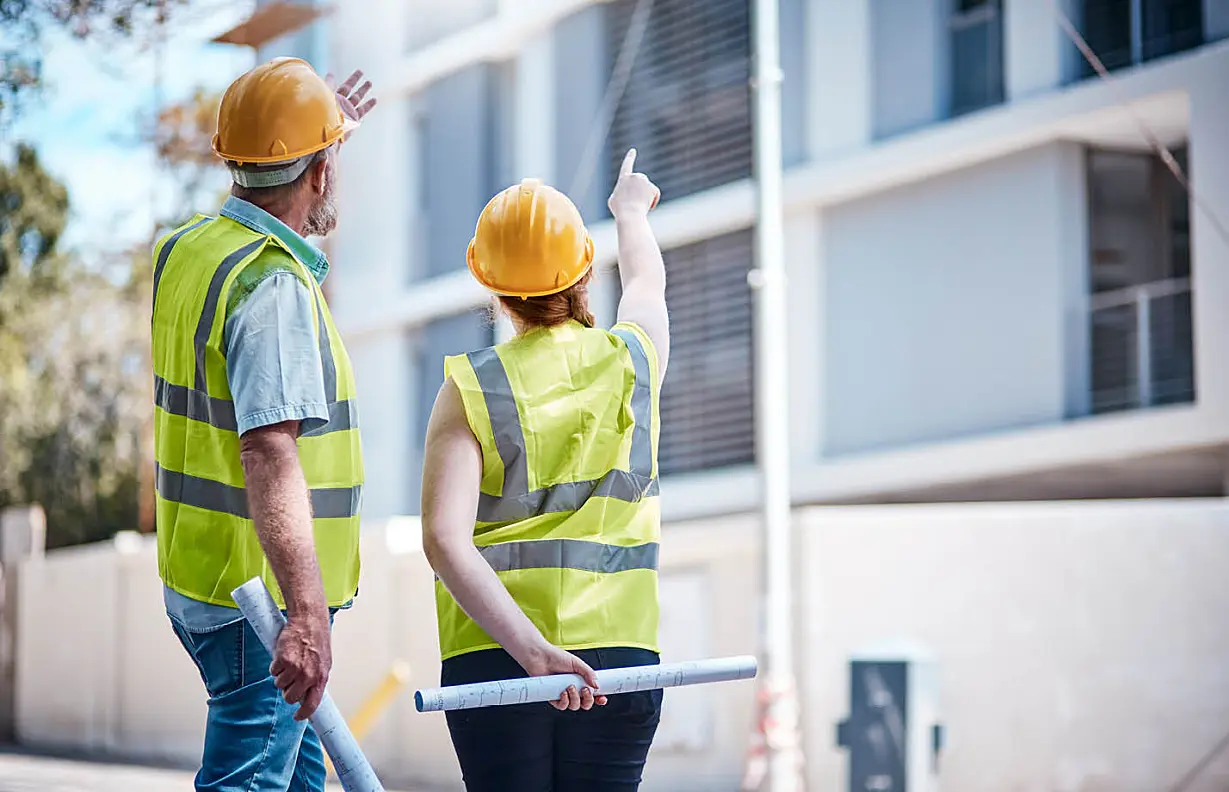 two engineers with yellow hard hats and rolled up plans, woman pointing to something in building
