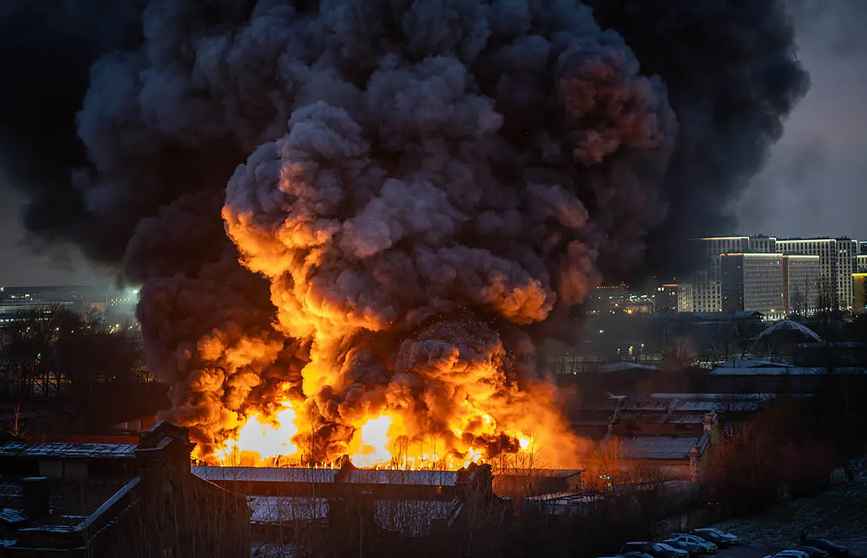 Massive fire engulfs industrial area at night, with large orange flames and thick black smoke.