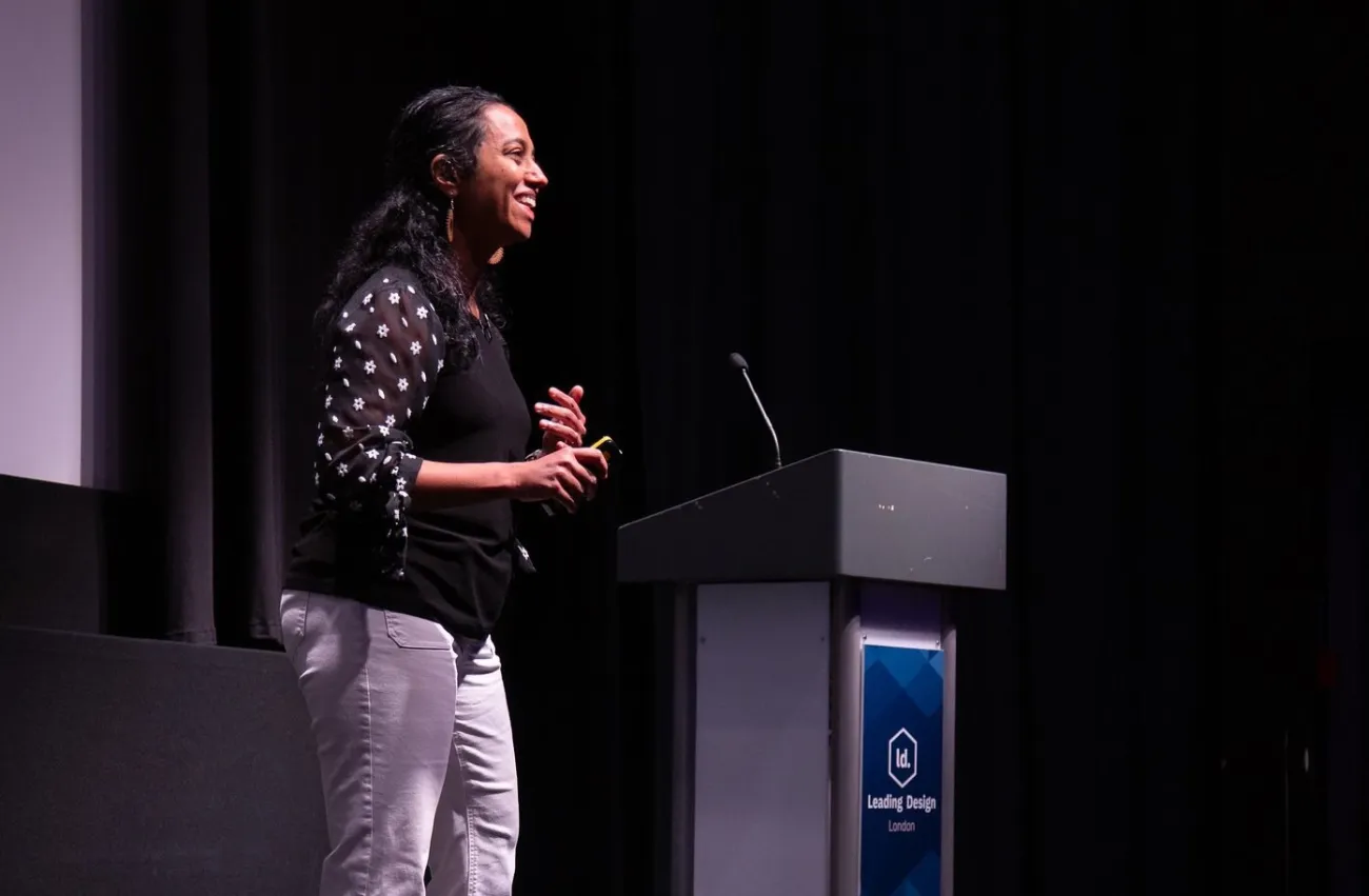 A woman stands on stage, smiling and speaking, beside a podium with a microphone. She wears a black and white polka dot top, light pants, and brown boots, in a dimly lit auditorium.