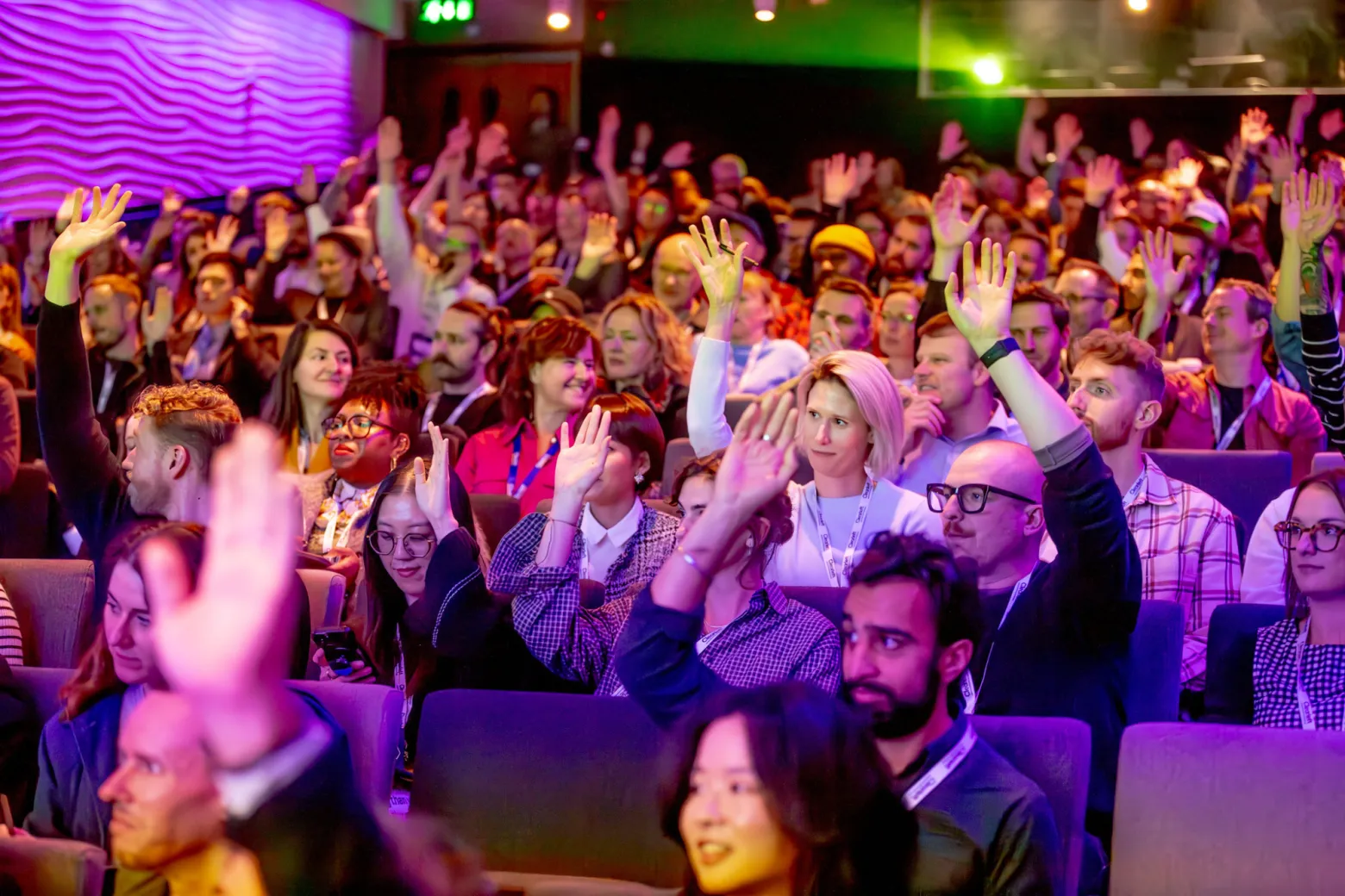 A large group of people sit in an auditorium, many with their hands raised, engaging in what appears to be a conference or seminar. The atmosphere is lively and colorful, with bright lights illuminating the crowd.