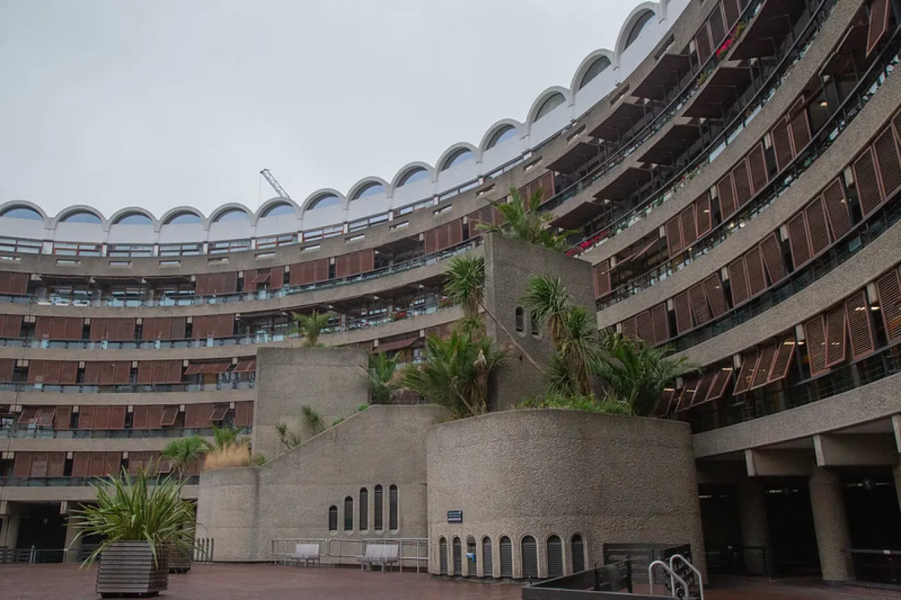 The Barbican Centre is pictured. A large, curved, multi-story concrete building with many windows and balconies, featuring planters with palm trees and a cloudy sky overhead.