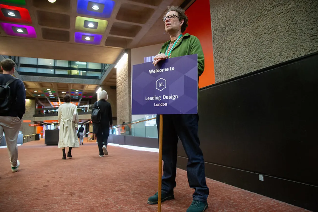 A man in a green jacket holds a purple sign reading ‘Welcome to Leading Design London’ in a modern indoor venue with colourful ceiling lights, as people walk past in the background.