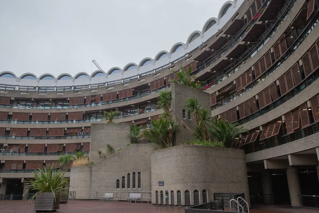 The Barbican Centre is pictured. A large, curved, multi-story concrete building with many windows and balconies, featuring planters with palm trees and a cloudy sky overhead.