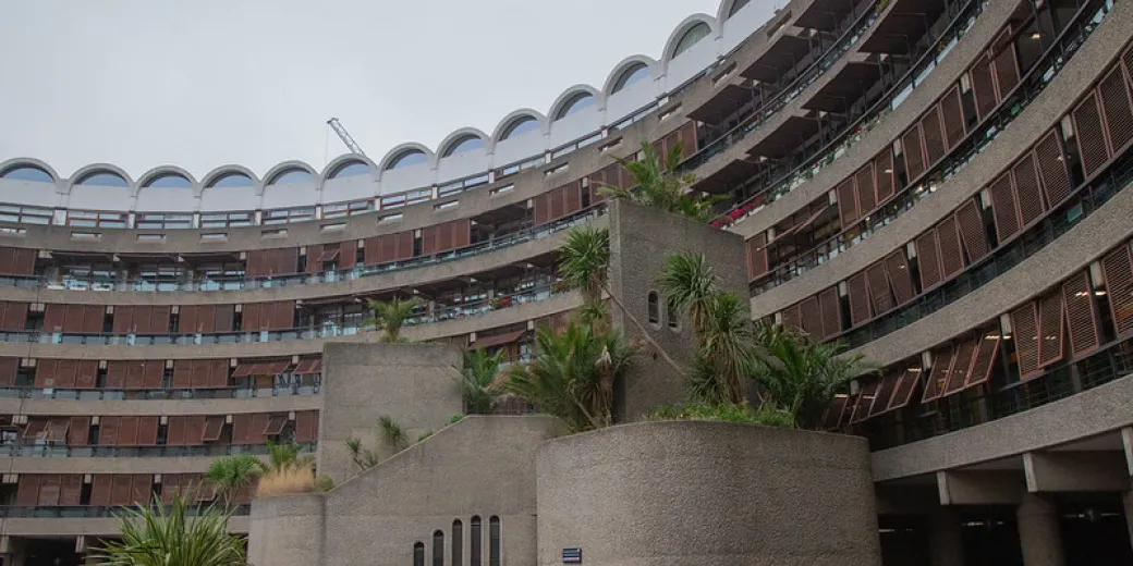 The Barbican Centre is pictured. A large, curved, multi-story concrete building with many windows and balconies, featuring planters with palm trees and a cloudy sky overhead.