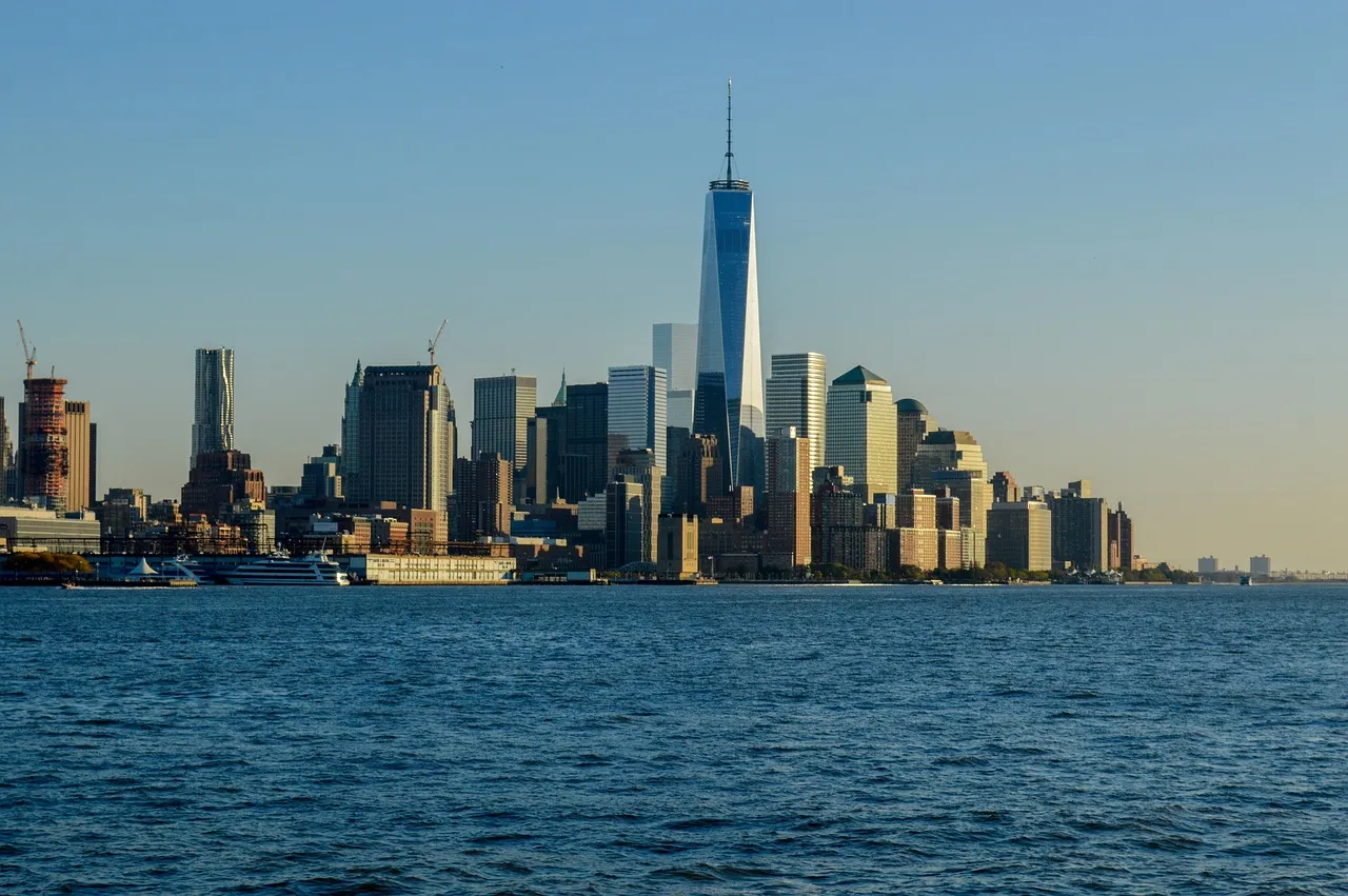 One World Trade Center rises above the Lower Manhattan skyline across a broad expanse of rippling blue water. Clear skies and warm light give the waterfront view a calm, open feel.