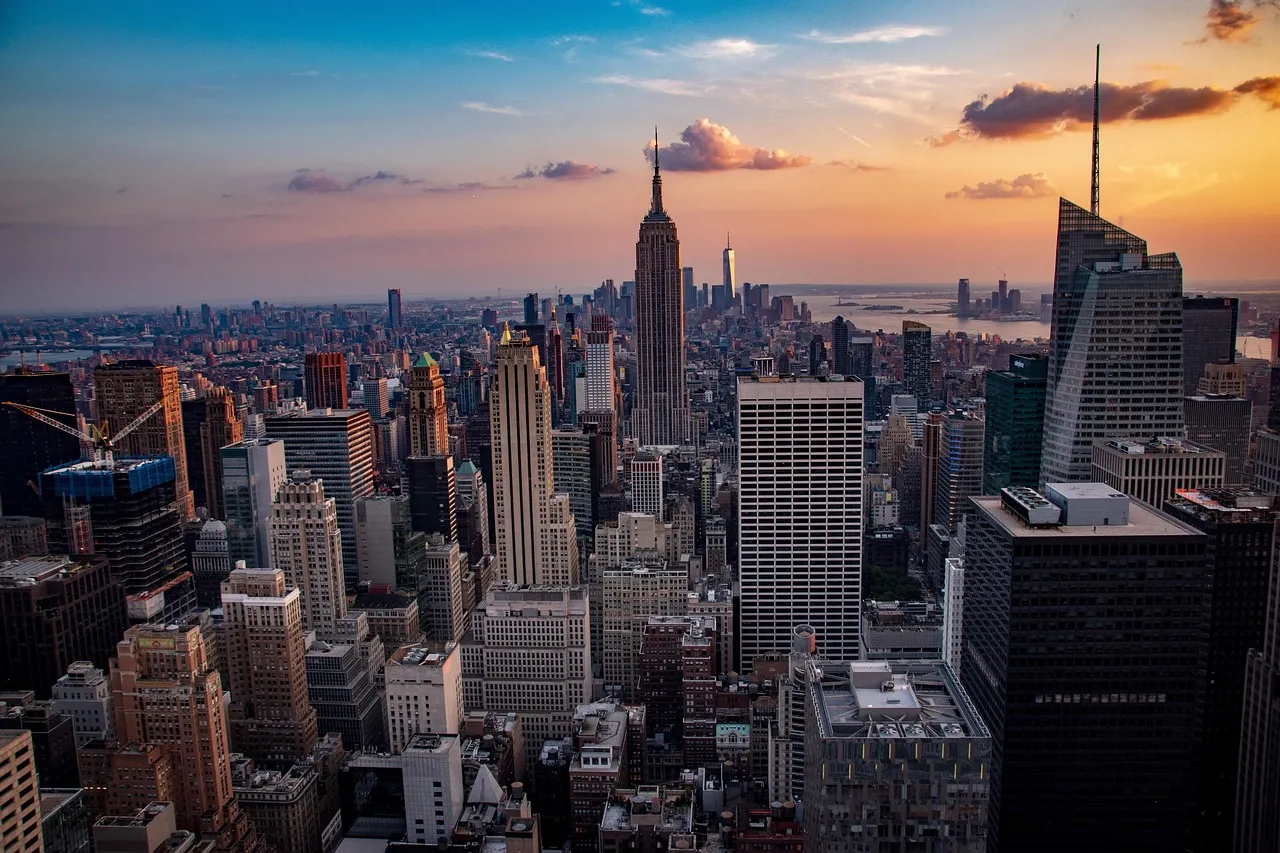 Manhattan's dense skyline surrounds the Empire State Building beneath a blue, pink, and orange sunset sky. The elevated view captures a dramatic, warmly lit cityscape.