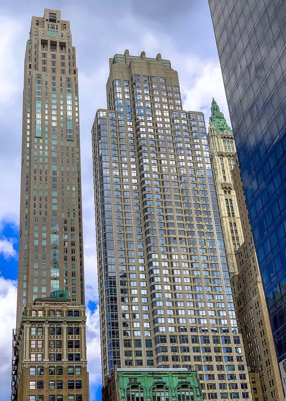 A street-level view looks sharply upward between tall New York skyscrapers, with stone facades, reflective windows, and an ornate green-roofed tower. Clouds and patches of blue sky emphasize the imposing height of the buildings.