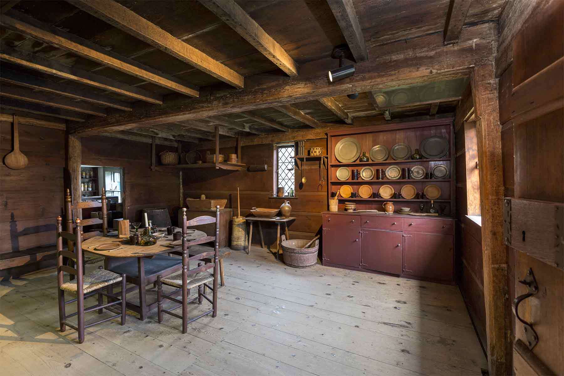 Inside the John Ward House. Wooden beams run the length of the ceiling. A wooden table and chairs sits to the side and on the back sits a cabinet displaying dishes.