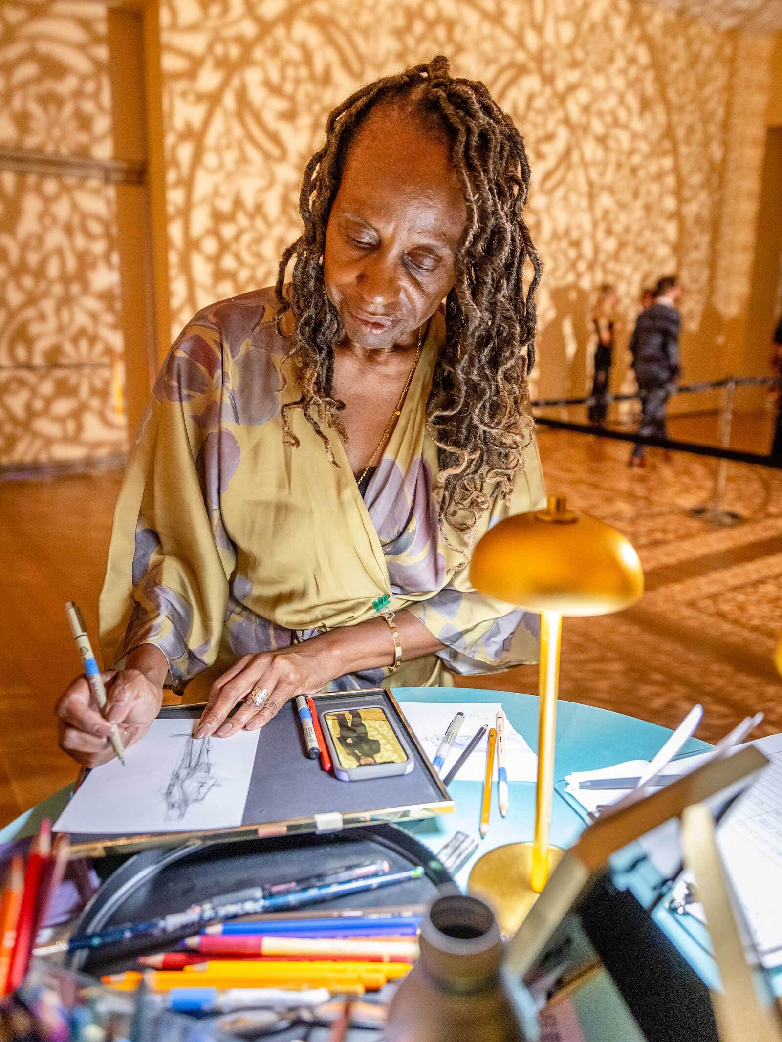 Rhonda Corley sits at a table in laid with colored pencils, quick-sketching a gala guest in a museum gallery.