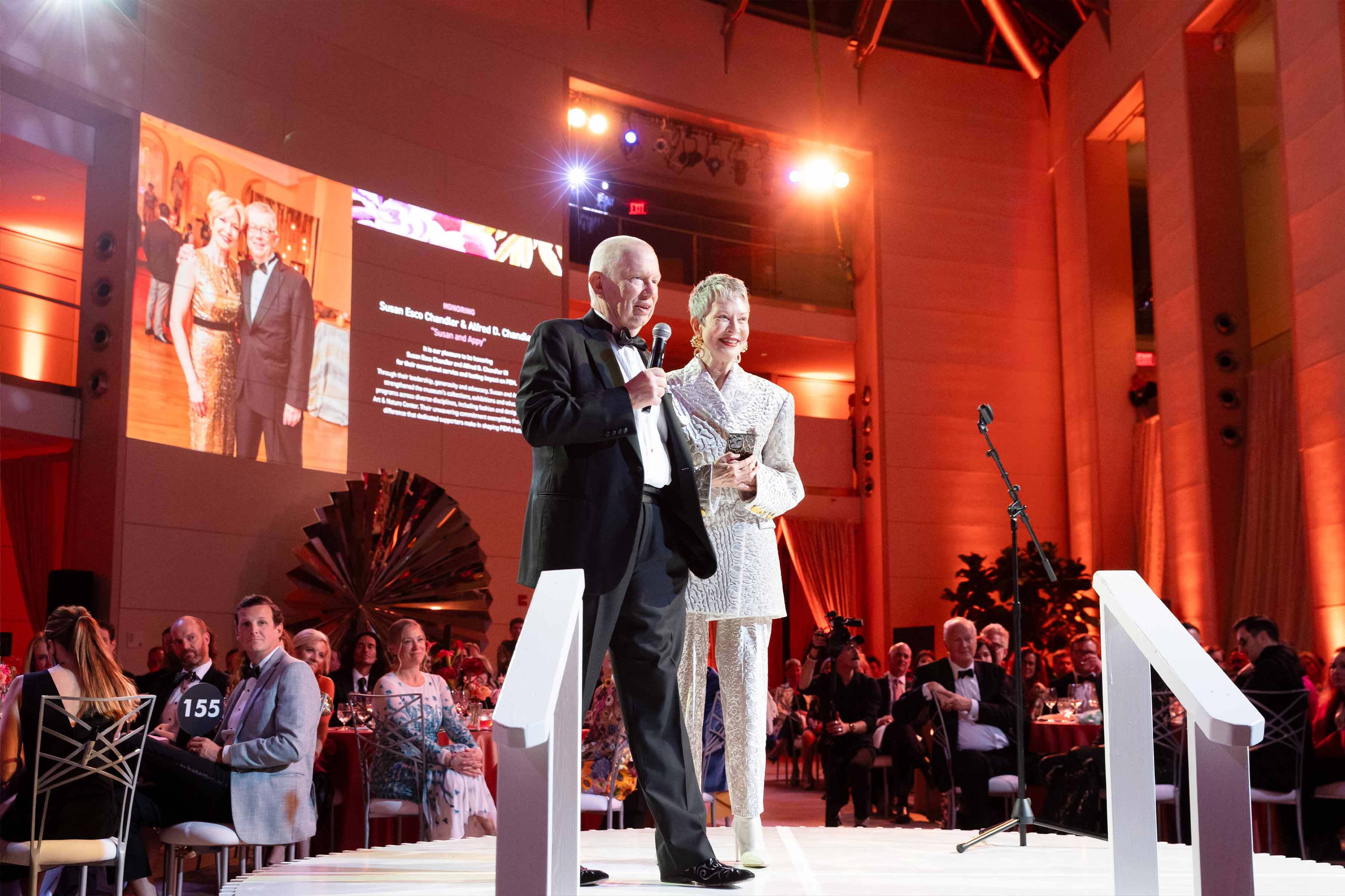 Susan and Appy Chandler, wearing tuxedoes, stand on a stage with microphones to accept their gala honors.