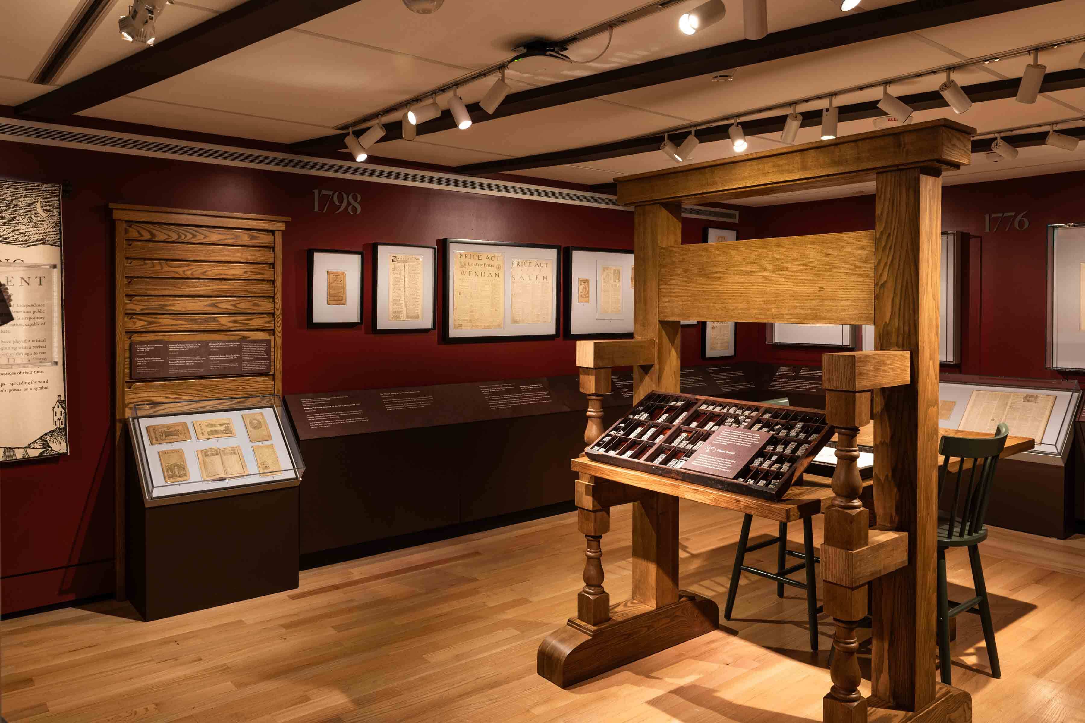 Museum gallery with a wooden historic printing press display, framed documents, and deep red walls under track lighting.