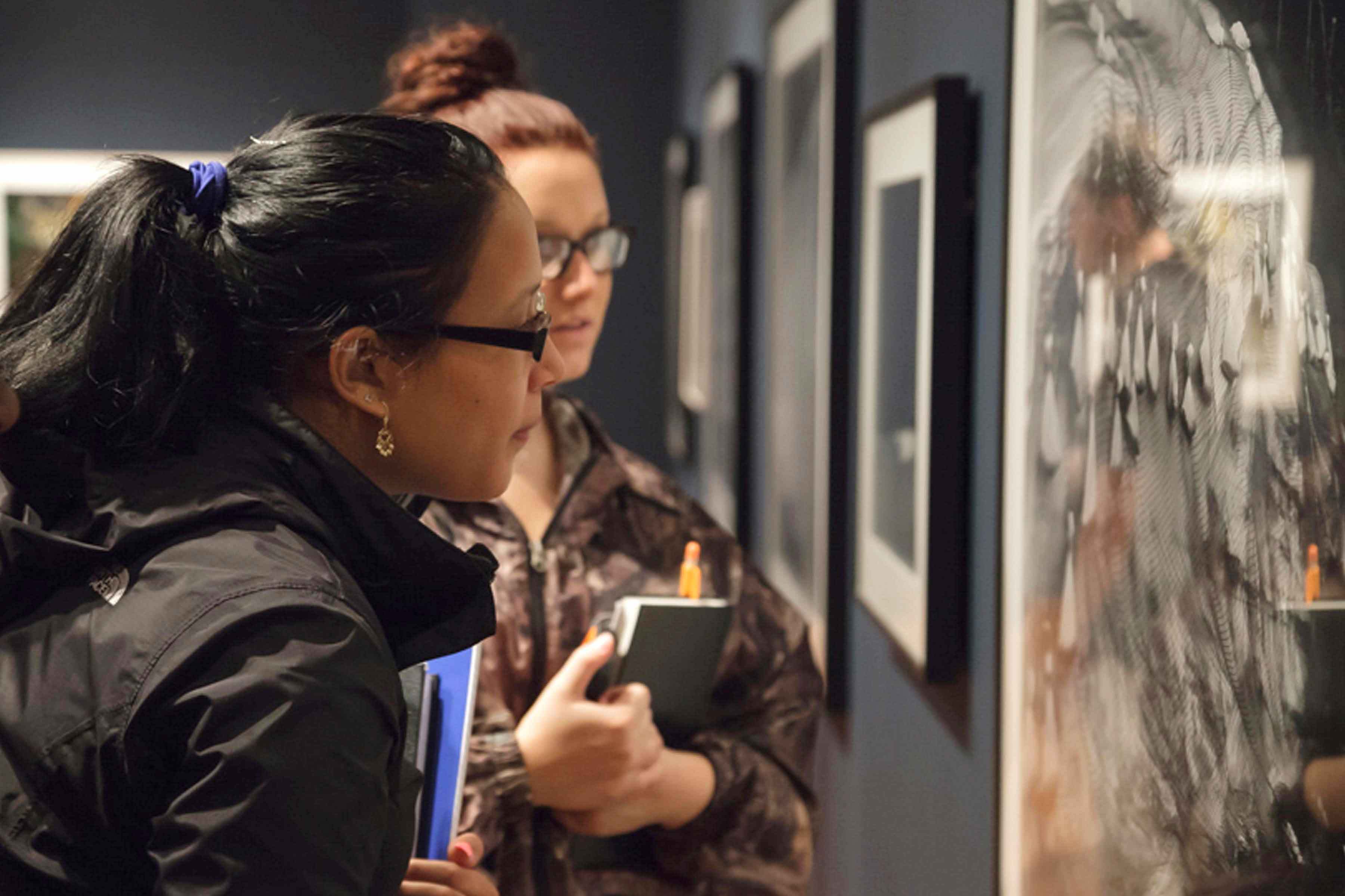 A couple of young women holding notebooks look at a photograph on a wall in the museum  Classroom Resources for Teachers