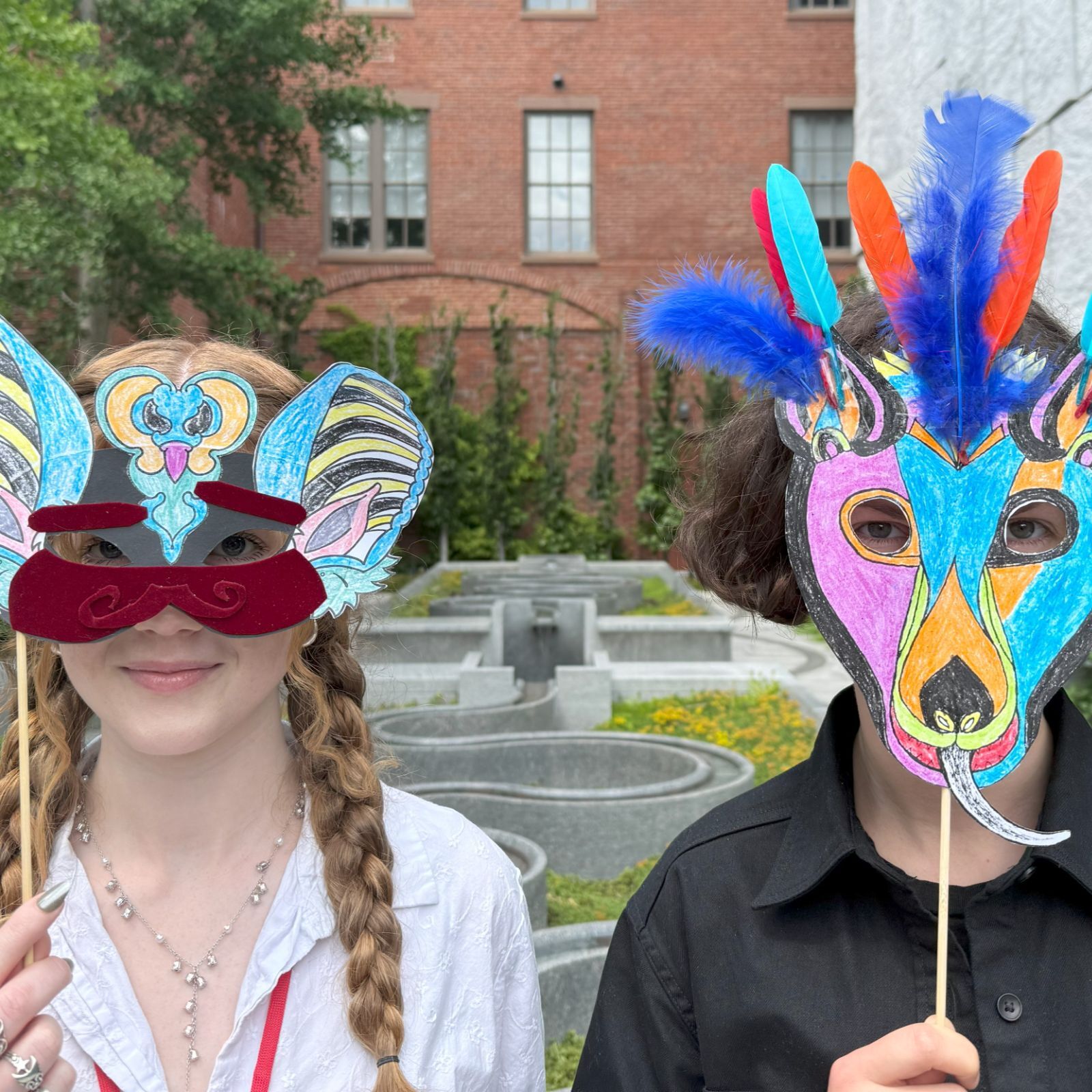 Two people pose outdoors holding colorful, hand-drawn paper masks on sticks.