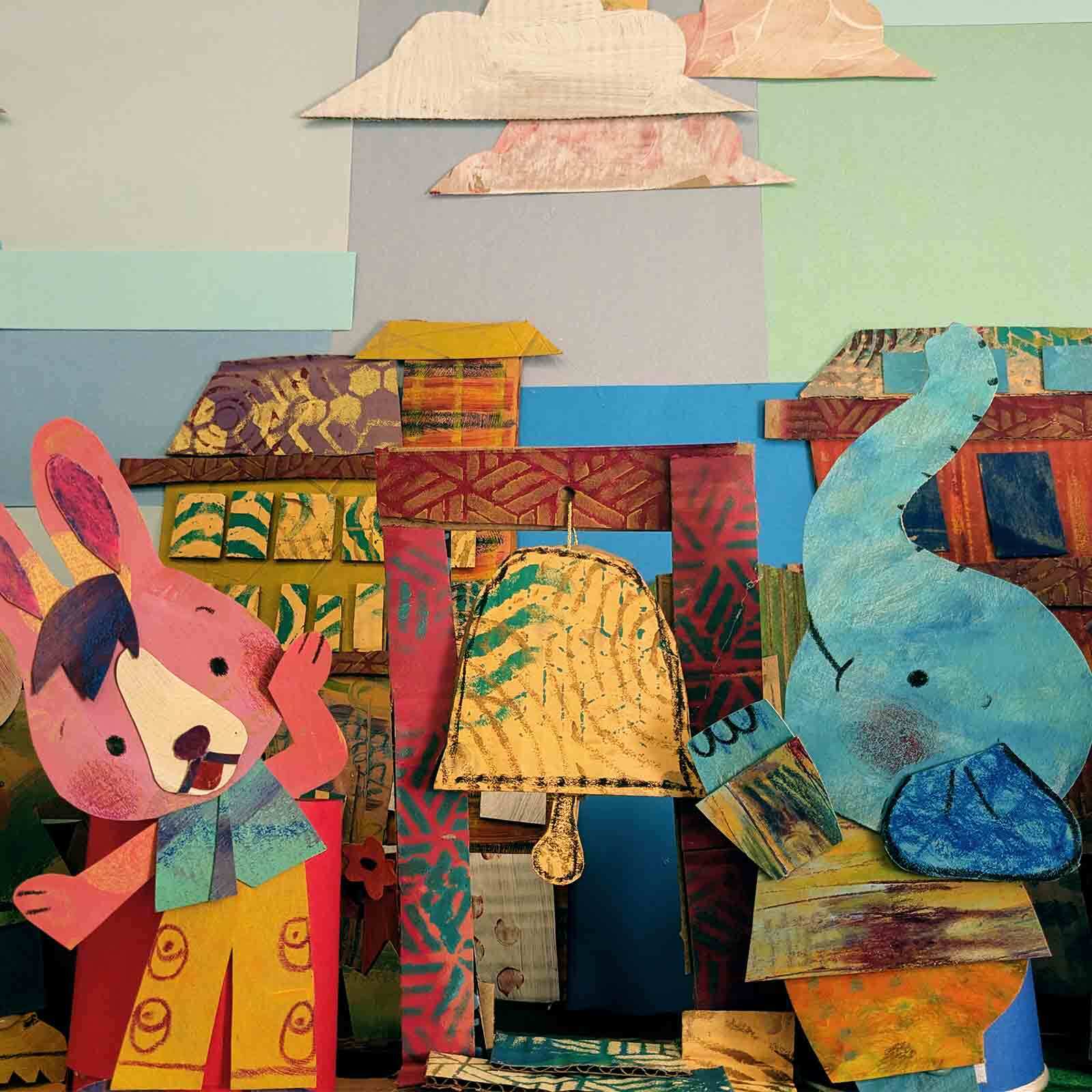 A pink bunny and blue elephant made out of painted paper stand besides a yellow paper bell