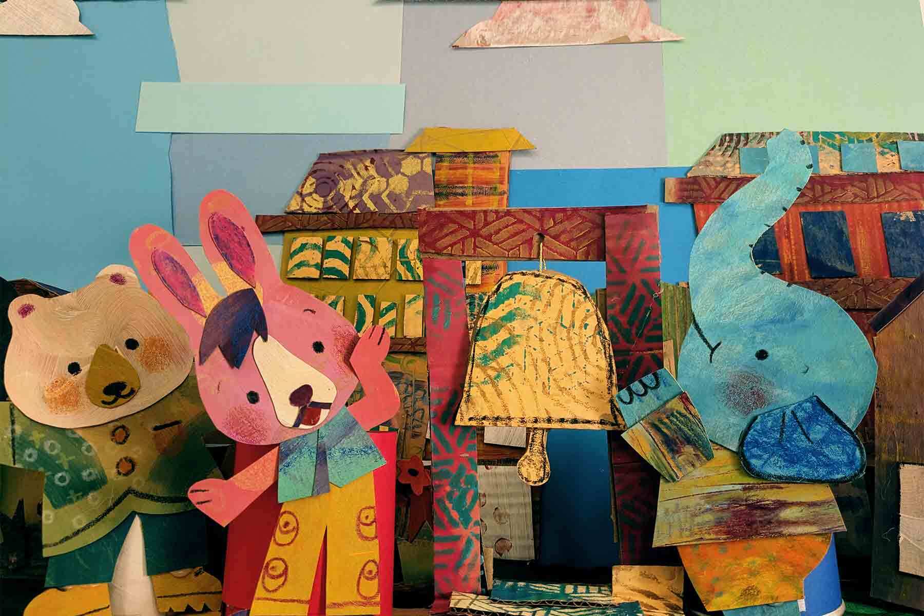 A pink bunny and blue elephant made out of painted paper stand besides a yellow paper bell