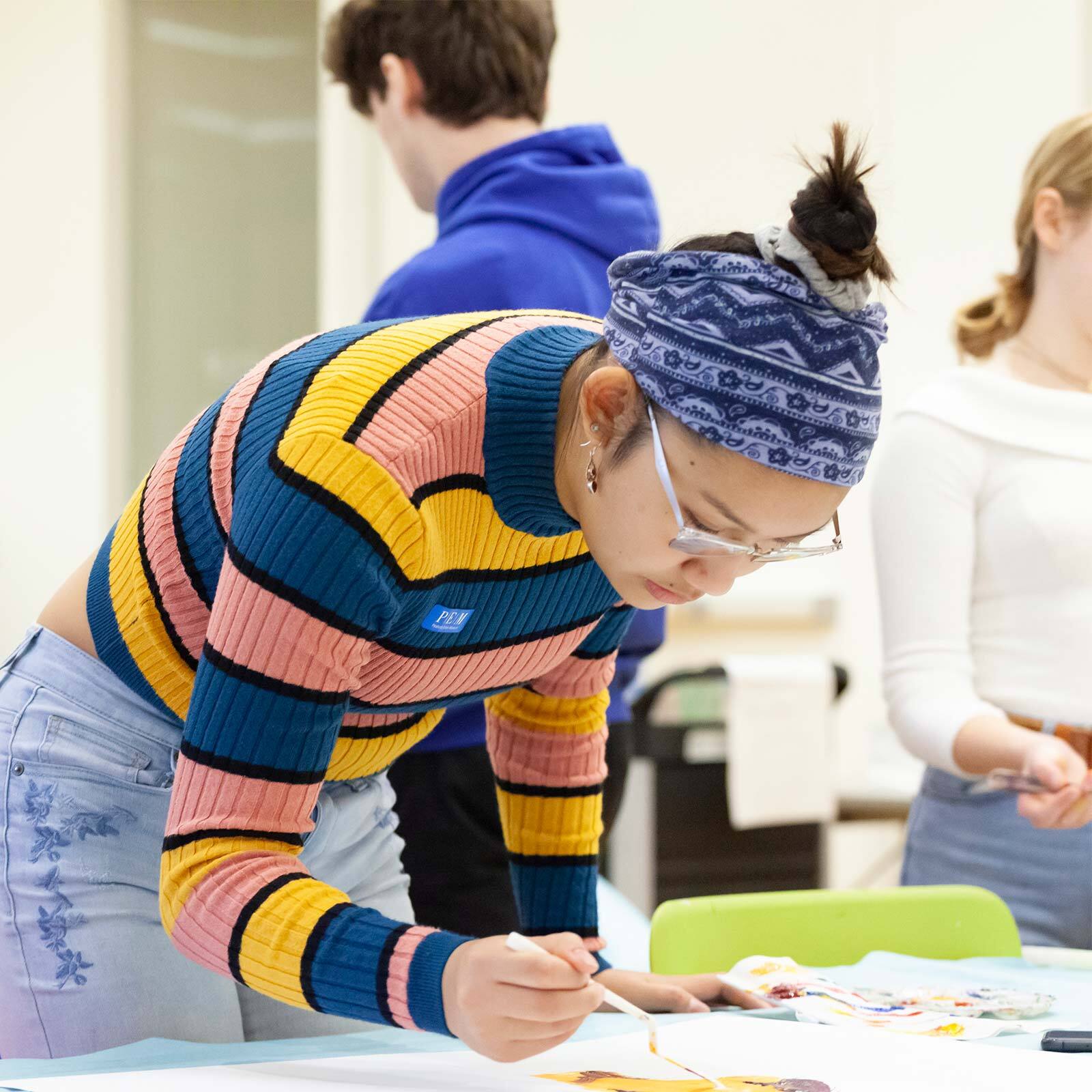 A person wearing a colorful striped sweater and headband paints on a large sheet of paper in a classroom filled with other students working on art projects.