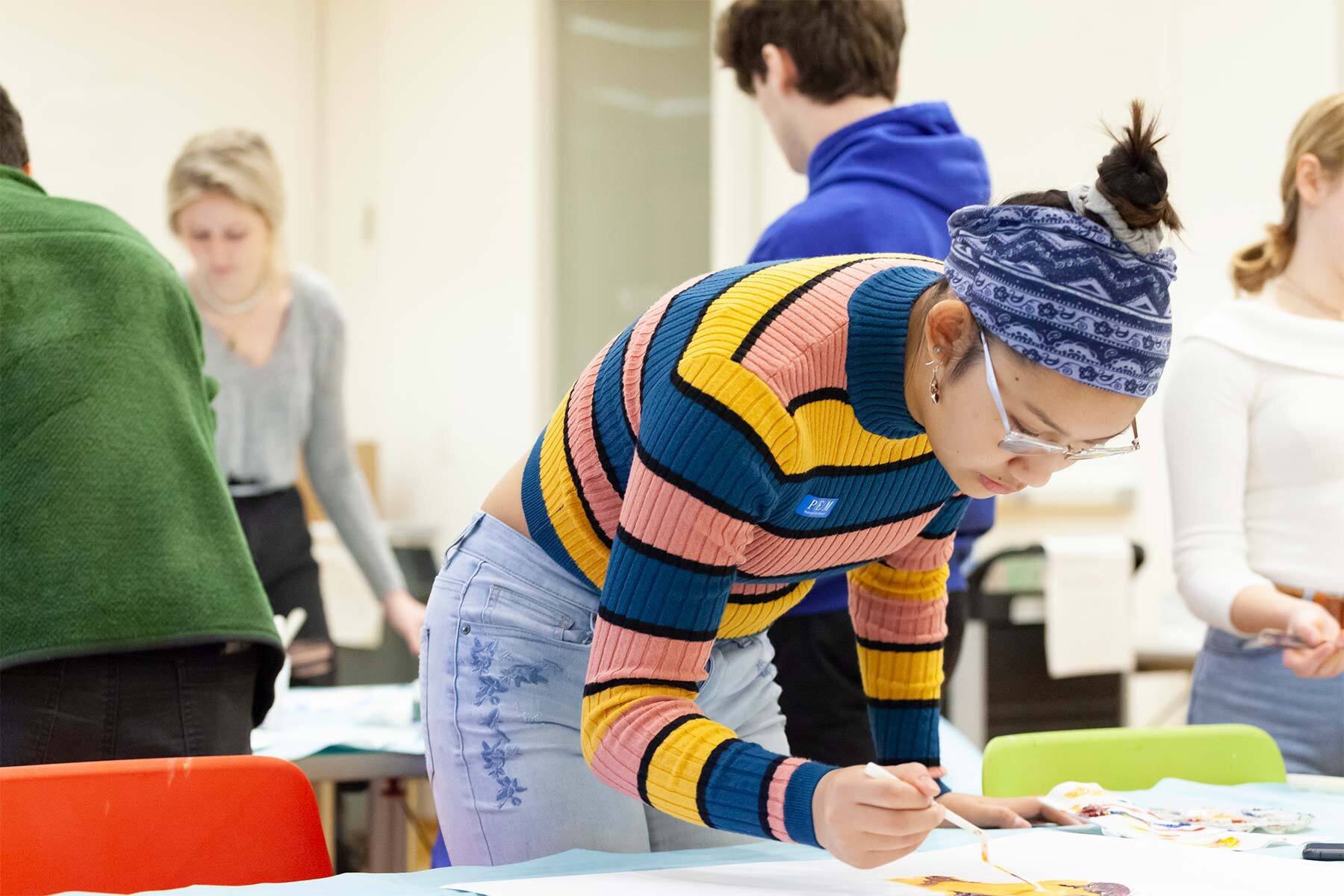 A person wearing a colorful striped sweater and headband paints on a large sheet of paper in a classroom filled with other students working on art projects.