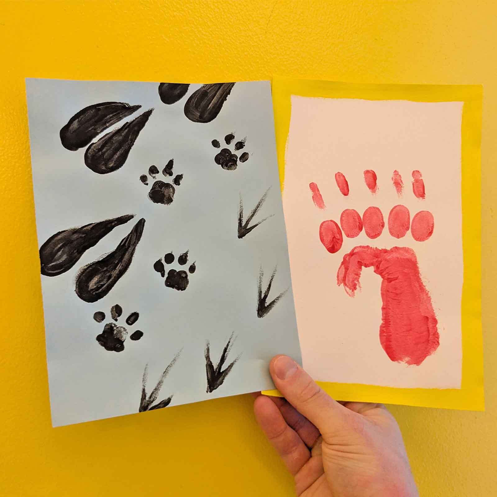 Handmade card with painted animal tracks and a red handprint on colorful paper against a yellow background.
